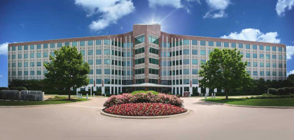 Modern glass-fronted office building centered behind roundabout flowerbed, trees, and clear blue sky
