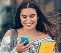 Young woman smiling while checking smartphone, holding textbooks outdoors