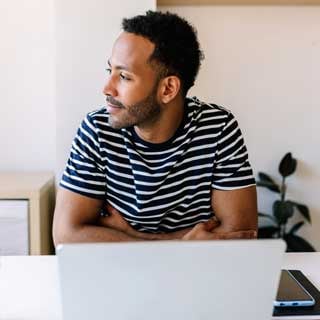 A man in a striped shirt sitting at a desk in front of a laptop, gazing off to the side.