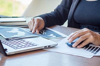 Person analyzing printed data charts beside laptop, pointing and using mouse