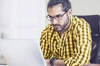 Young man wearing glasses and yellow plaid shirt concentrating on laptop at office desk