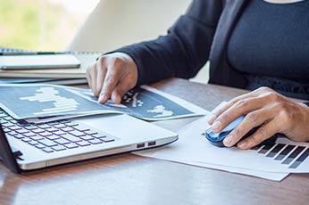 Person analyzing printed data charts beside laptop, pointing and using mouse