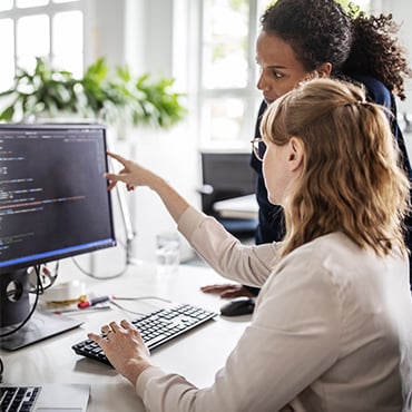 Developer pointing at code on monitor while mentoring colleague in bright open office