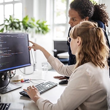 Woman points at code on monitor while colleague types in bright office