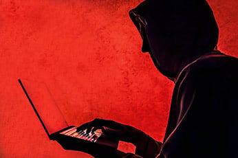 Hooded figure typing on laptop against ominous red background, suggesting cybercrime activity