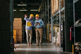 Two warehouse workers wearing hard hats inspecting inventory while walking an aisle