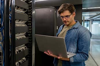 IT technician wearing safety glasses inspecting server rack while using laptop