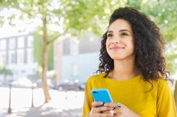 Smiling woman using smartphone while standing outdoors on sunny street