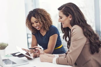 Two colleagues smiling and discussing documents on a tablet at a bright office desk