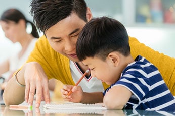 Parent guiding young boy as they write in a notebook at kitchen table