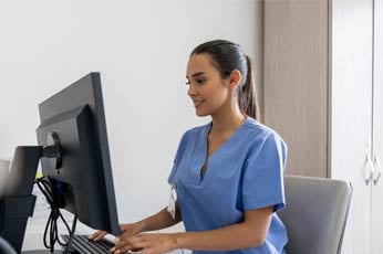 Medical nurse in blue scrubs typing on desktop computer at clinic reception desk