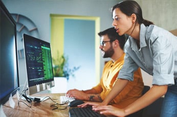 Woman leaning over while coworker codes at dual monitors in modern office