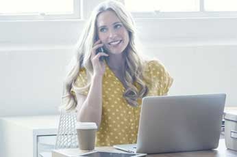 Smiling woman talking on phone while working at laptop in bright office