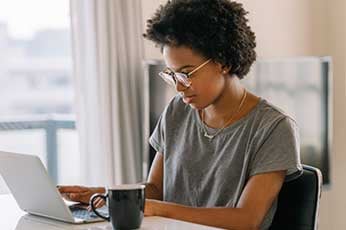 A woman wearing glasses sitting in front of a window at a desk and using a laptop.