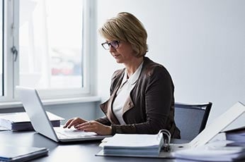 Mature woman typing on laptop at organized office desk with binders and paperwork