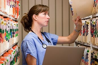 Nurse in scrubs reviewing patient paper charts in filing room with laptop