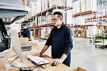 Warehouse worker using desktop computer at packing station amid shelving