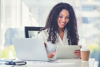 Woman using tablet and laptop at bright office desk, smiling during a remote meeting