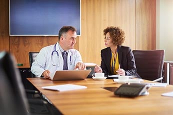 Doctor and businesswoman discussing patient case over laptop in conference room