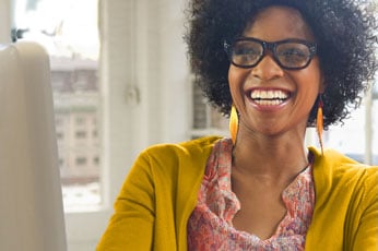 Smiling woman wearing glasses and yellow cardigan, working on laptop by bright window