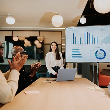 Woman presenting performance charts on screen while colleagues applaud in conference room