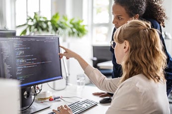 Senior developer pointing at code on monitor while colleague types
