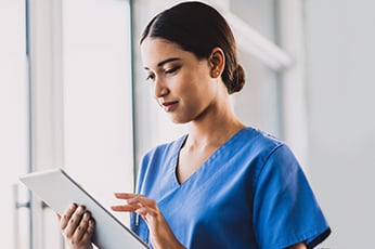 Nurse in blue scrubs reviewing patient information on a tablet beside a bright window
