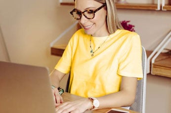 Smiling young woman in yellow shirt typing on laptop at tidy home desk