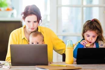 Mother multitasking on laptop while baby watches and daughter uses tablet at kitchen table