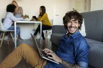 Smiling man using laptop on floor while colleagues chat at table