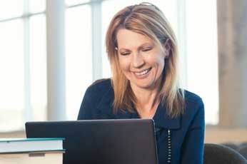 Middle-aged woman smiling while working on laptop near stacked books in sunlit office window