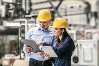 Male and female engineers in yellow hard hats reviewing clipboards on factory floor