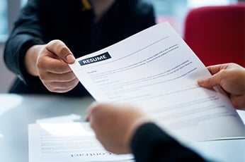 Person handing a paper labeled "RESUME" to interviewer across table
