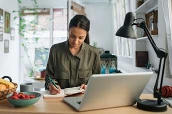 Young woman writing in notebook at tidy home desk with open laptop and lamp