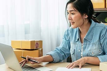 Woman reviewing online orders on laptop while pointing with pen surrounded by shipping boxes