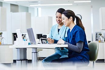 Two nurses reviewing patient records at clinic workstation, smiling and discussing