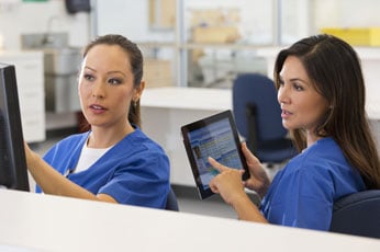 Two nurses reviewing patient chart on tablet while discussing care at busy nursing station
