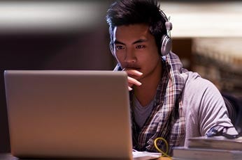 College student wearing headphones intently working on laptop in cozy study area