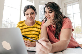 Adult pointing at laptop screen while studying with teen in sunlit room