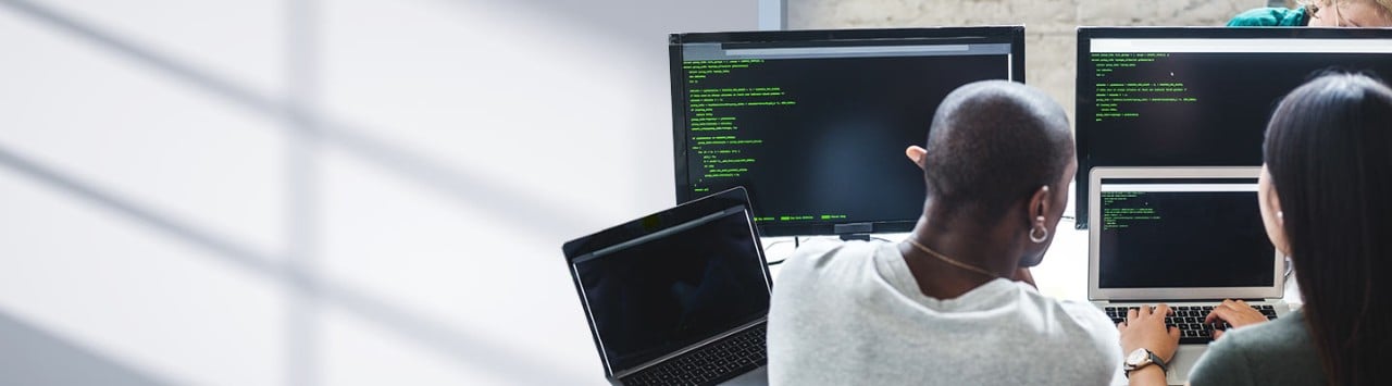 Two programmers collaborating at laptops and monitors reviewing green terminal code