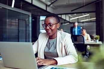 Smiling professional woman typing on laptop at modern open-plan office desk, colleagues blurred behind