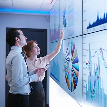 Two office professionals pointing to interactive wall of colorful pie charts and time-series graphs