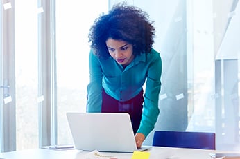 Woman leaning over laptop, focused on work in sunlit office