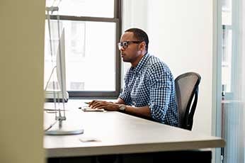Man in checkered shirt typing at desktop computer in bright office