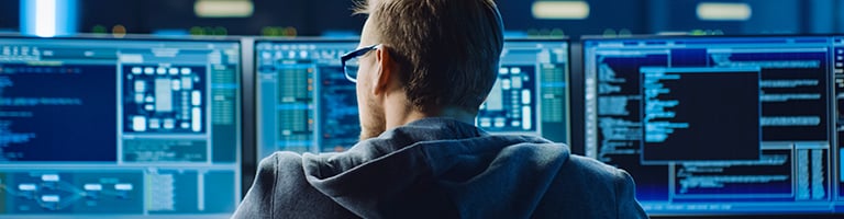 Software engineer wearing glasses and hoodie monitors multiple screens with code