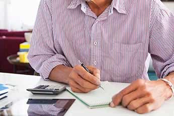 Person writing notes in a notebook at desk with calculator and tablet