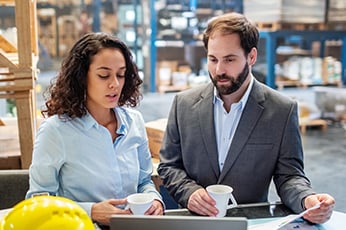 Two colleagues holding coffee reviewing laptop and documents in warehouse