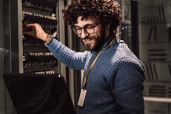 Smiling IT technician with curly hair and glasses configuring server rack with laptop