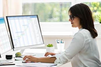 Young professional woman typing on keyboard at dual monitors in sunlit office