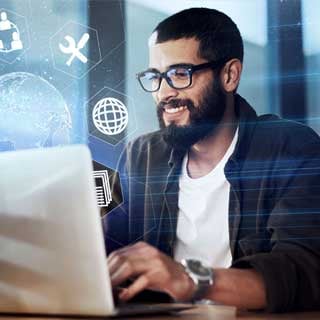 A smiling man with a beard and glasses working on a laptop surrounded by digital icons, representing innovation and AI education in a modern learning environment.